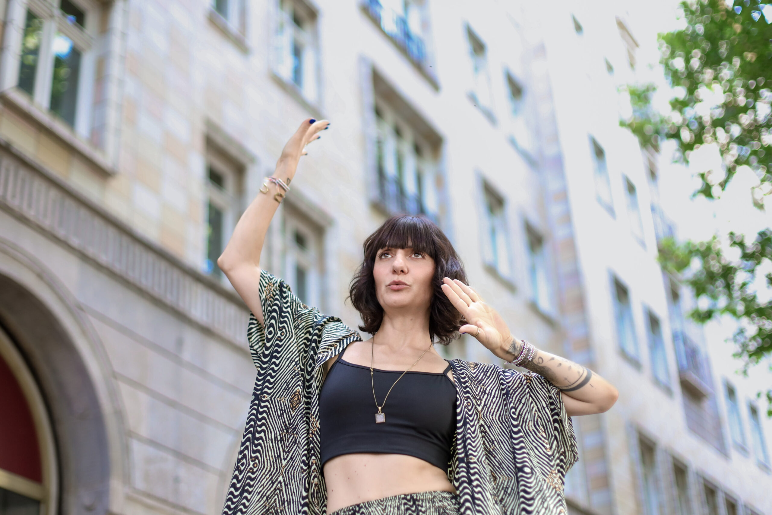 Captured from a low angle, Nadia El-Nadi, founder of Higher Resolution, stands with her body facing more frontally, arms and hands raised in expressive movements, with her mouth shaped as if whistling. She gazes into the distance, symbolizing anticipation and strength, against the backdrop of a massive, old building. The image conveys a powerful readiness to face what’s ahead in an urban setting.